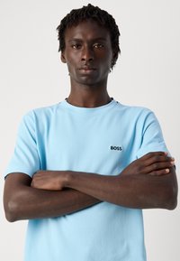 Gaiši zils īsām piedurknēm T-krekls ar apaļu kakla izgriezumu un melnu "BOSS" logo kreisajā krūtī. Materiāls izskatās teksturēts. Modelis ar saslēgtām rokām.
