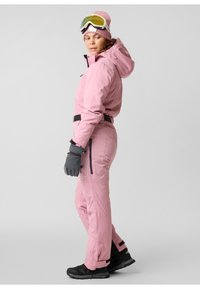 Pink winter suit with a hood, black belt, and pockets. Grey gloves and black boots complete the outfit. Ski goggles rest on the forehead.