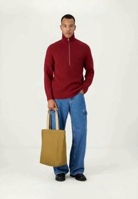 Roter gestrickter Pullover mit hohem Kragen und halbem Reißverschluss, kombiniert mit hellblauen, weit geschnittenen Jeans; hält eine beigefarbene Tragetasche aus Canvas.