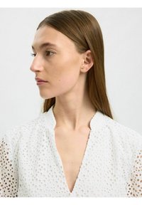 Vit blus i perforerat tyg med djup V-ringning och stående krage. Har texturerade hål genom hela designen.