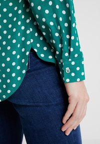 Blouse verte à pois avec une texture douce, présentant des pois blancs et un poignet boutonné. Associée à un jean en denim foncé.