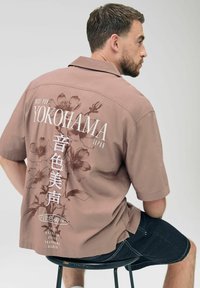 Košulja kratkih rukava u svijetlosmeđoj boji s cvjetnim i tekstualnim grafikonima na leđima; karakterizira je opušteni kroj i meka tekstura tkanine.