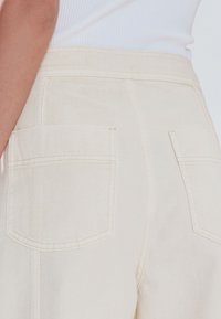 Un pantalon en coton beige avec des poches arrière, une texture lisse et des coutures visibles le long de la ceinture et des poches.