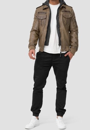 Homme portant une veste en cuir marron par-dessus un sweat à capuche gris, un t-shirt blanc, un pantalon de jogging noir et des baskets blanches, debout sur un fond gris.