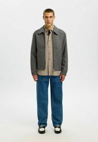 Siva jakna iz volnene mešanice s ovratnikom, bež srajca s petimi gumbi pod njo, ohlapne modre jeans hlače in črno-beš čevlji.