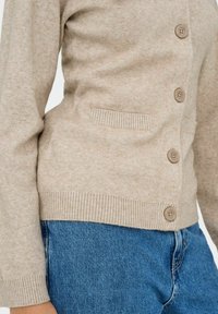 Cardigan beige avec une texture douce, cinq boutons et des poignets côtelés. Comprend des poches latérales et une silhouette ajustée, associé à un jean bleu.