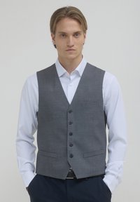 Junger Mann mit hellbraunen Haaren, der eine graue, geknöpfte Weste über einem weißen Hemd und dunklen Hosen trägt, vor einem hellen, einfarbigen Hintergrund.