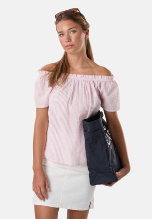 Jeune femme aux longs cheveux tient un sac cabas bleu marine, porte un haut rose clair épaules dénudées et une jupe blanche, avec des lunettes de soleil sur la tête.