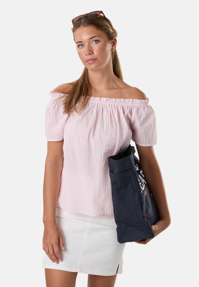 Jeune femme aux longs cheveux tient un sac cabas bleu marine, porte un haut rose clair épaules dénudées et une jupe blanche, avec des lunettes de soleil sur la tête.