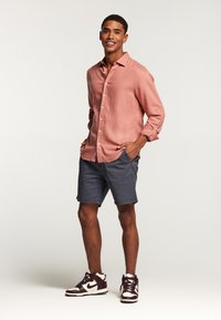 Chemise boutonnée à manches longues rose clair, short gris et baskets bordeaux. La chemise a une coupe décontractée et des poignets retroussés avec une légère texture.