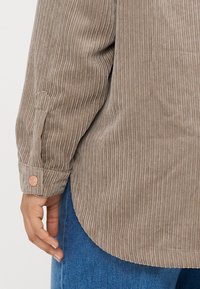 Rudas korduroyo marškiniai su vertikaliu šonkauliu tekstūra, mygtukų rankogaliai ir išlenktas kraštas. Dėvima su mėlynomis džinsomis.