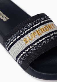 Chausson de bain marine avec une lanière tissée ornée de motifs blancs et bleu clair, accentué par un logo doré brodé. Semelle texturée.