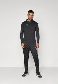 Juoda sportinė apranga, susidedanti iš ilgomis rankovėmis su užtrauktuku kaklo srityje, mėlyna logotipu ir raštuotomis rankovėmis, derinama su juodomis, siaurėjančiomis kelnėmis ir sportiniais batais.