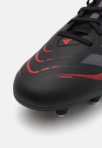 Botín de fútbol negro con parte superior sintética texturizada, con acentos rojos y un diseño de línea curva. Incluye tapones moldeados para tracción.