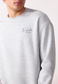 Grå hettegenser laget av mykt stoff med rund hals. Har en marineblå logo med teksten "The Sailor" på venstre brystområde.