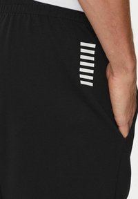 Pantaloni sportivi neri realizzati in tessuto morbido, con una tasca laterale e strisce orizzontali bianche come dettaglio di design. Vita elastica visibile.