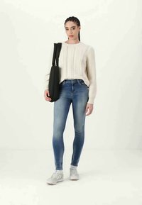 Suéter de malha creme com relevo, jeans skinny azuis e ténis brancos. Saco preto atravessado no ombro. Fundo neutro.