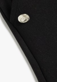 Zwarte stof met een gladde textuur, met een ronde zilveren knop met een gestructureerde rand en embleemdetail aan de bovenrand.