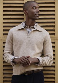 Beige wool sweater with a V-neck and a subtle texture, layered over a checked shirt with blue and white patterns. Dark corduroy pants and brown belt.