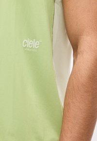 Πράσινη και λευκή αμάνικη αθλητική μπλούζα με το λογότυπο "ciele athletics" στο στήθος, δίπλα σε ακάλυπτο χέρι.