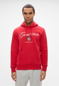 Jeune homme portant un sweat à capuche rouge avec "Gant 1949" et un logo de blason, associé à un pantalon de survêtement gris clair, se tenant devant un fond uni.