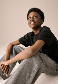 Zwart katoenen T-shirt gecombineerd met lichtgrijze, losvallende spijkerbroek. De achtergrond is neutraal, wat het contrast van de outfit versterkt.