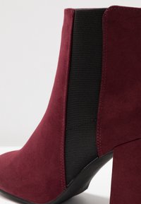 Botín de ante burdeos con panel elástico lateral negro y tacón grueso de altura media sobre fondo blanco.