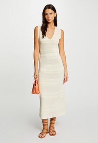 Strickmidi-Kleid in cremiger Farbe mit V-Ausschnitt, gewellten Kanten und strukturierten Mustern. Kombiniert mit einer rot-orangefarbenen Handtasche und braunen Riemchensandalen.