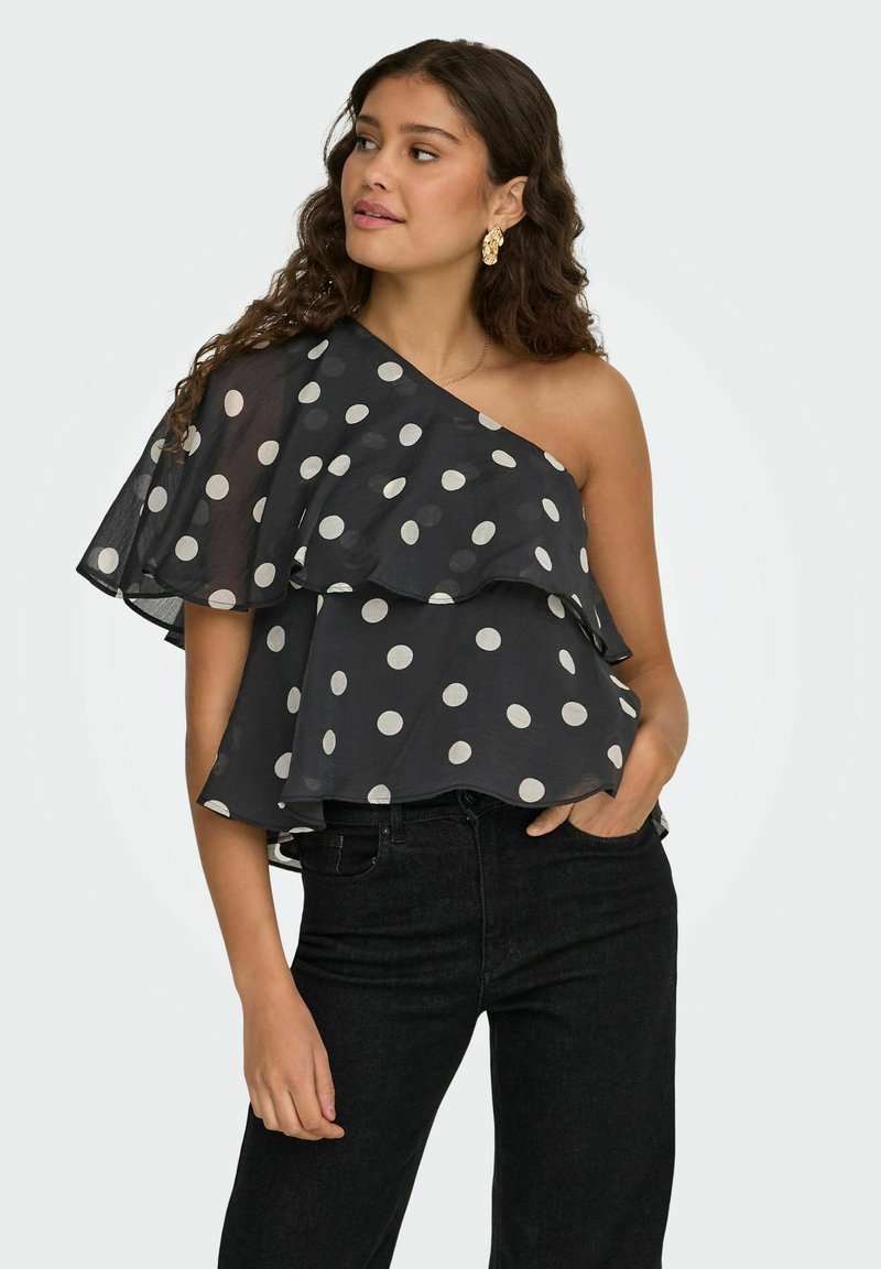 Femme portant un haut noir à une épaule avec des volants et des pois blancs, ainsi qu'un pantalon noir, posant avec une main dans la poche.