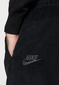 Černé kordové jogger kalhoty s kapsou, zdobené vyšitým stříbrným logem Nike. Tkanina vykazuje výrazné vertikální pruhy a texturu.