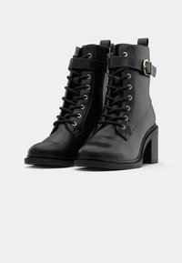 Paire de bottines en cuir noir à talons épais, avec lacets à l'avant et sangles à boucle sur un fond clair uni.