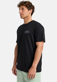 Sort bomulds-T-shirt med rund halsudskæring og korte ærmer, prydet med et lille broderet logo på brystet i en kontrasterende farve.