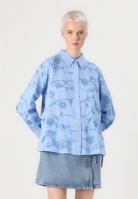 Svetlo modra bombažna srajca s cvetličnim vezenjem, z gumbi spredaj, s koničastim ovratnikom in dolgimi rokavi. V kombinaciji z jeans krilom.