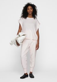 Hellrosa, lässig geschnittenes Oberteil mit hochgekrempelten Ärmeln, kombiniert mit schmal zulaufenden Hosen. Das Modell hält eine weiße Tasche. Schwarze Schuhe runden das Outfit ab.