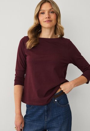 Kvinde med bølget blondt hår iført en langærmet, vinrød top og blå jeans, med den ene hånd, der justerer hendes linning.