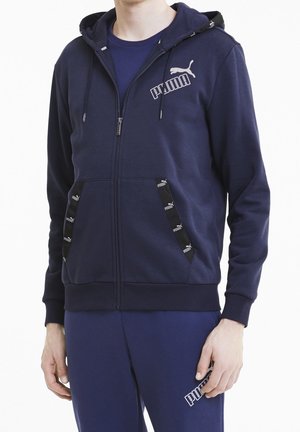 Mann trägt eine marineblaue Puma-Zip-Hoodie mit Logo auf der Brust und Taschenrändern, kombiniert mit passenden marineblauen Jogginghosen.