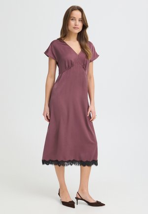 Kobieta stoi, ubrana w mauve midi sukienkę z czarną koronkową lamówką i brązowe szpilki typu slingback na jednolitym białym tle.