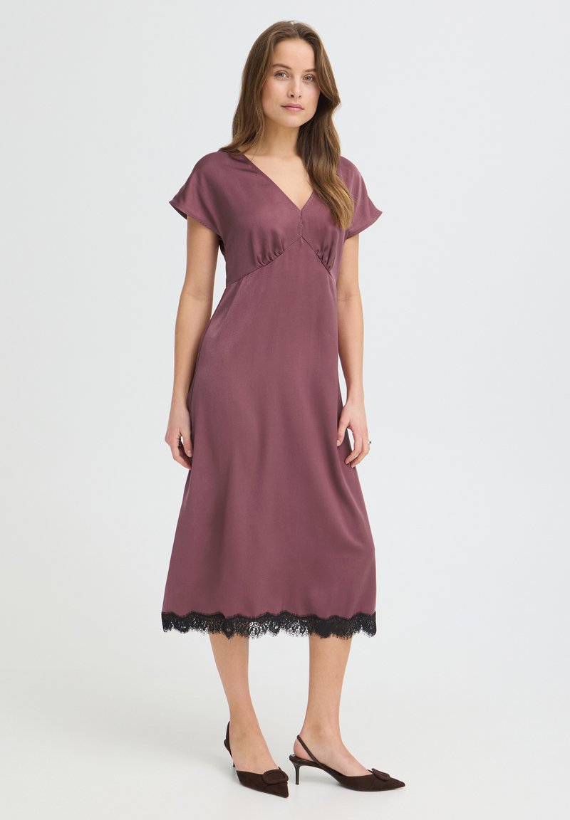 Femme debout portant une robe midi mauve avec une bordure en dentelle noire et des talons slingback marron sur un fond blanc uni.