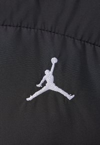 Fekete szövet, sima textúrával, amelyen egy fehér hímzett Jumpman logó található, jól láthatóan elhelyezve az anyagon.