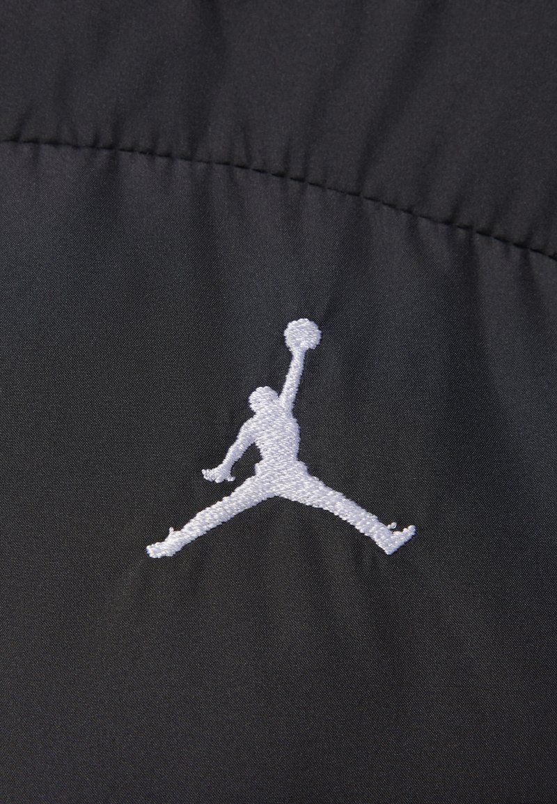 Czarna tkanina o gładkiej fakturze z białym haftowanym logo Jumpman, umieszczonym w widocznym miejscu na materiale.