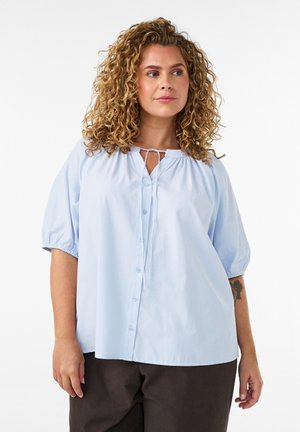 Femme aux cheveux bouclés portant un chemisier bleu clair boutonné avec des manches bouffantes et un pantalon noir, debout devant un fond blanc.