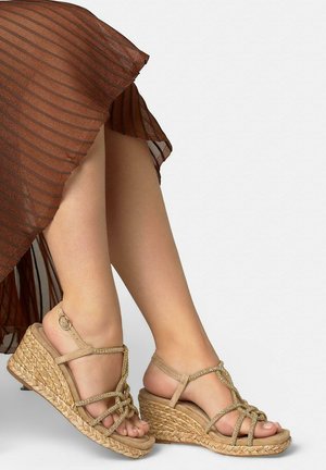 Sandalias beige de cuña trenzadas con tiras de cristal, combinadas con una falda marrón plisada, mostrando las piernas cruzadas sobre un fondo blanco.