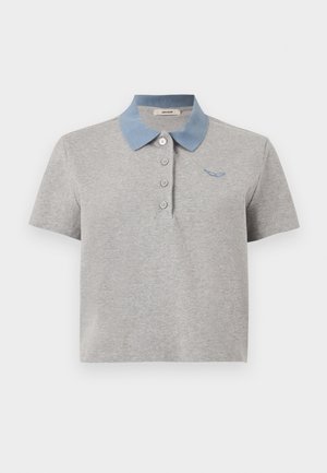 Femme portant un polo gris à manches courtes, un jean bleu à jambes larges avec fentes latérales, et des baskets marron avec des lacets rouges, debout devant un fond uni.