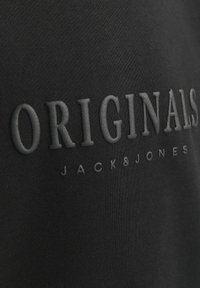 Svart bomullstyg med präglad text "ORIGINALS JACK&JONES" i fet stil, med en slät yta och subtil glans.