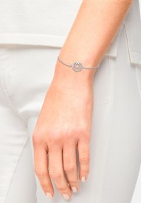Bracciale in argento con un ciondolo circolare a forma di cuore, incastonato con piccole pietre, su una delicata catena, indossato su un polso di carnagione chiara.
