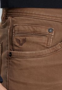 Ruske denim hlače s žepom, ki ima izvezeno logotip orla in črne kovinske klinčke, oblečene s temno modrim zgornjim delom.
