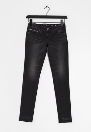 Schwarze Slim-Fit-Jeans mit Reißverschlusstasche und Knopfverschluss, die an einem schwarzen Bügel vor weißem Hintergrund hängen.