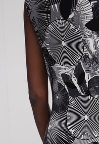 Närbild av en persons arm och torso som bär ett ärmlöst plagg i svart och vitt med cirkulära och bladliknande mönster.