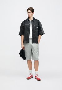 Νέος άνδρας που φορεί μαύρο κοντομάνικο πουκάμισο από τζιν, λευκό μπλουζάκι, γκρι σορτς, κόκκινα αθλητικά παπούτσια και κρατάει ένα μαύρο καπέλο, στέκεται μπροστά σε λευκό φόντο.