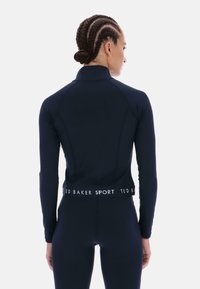 Námornícky atletický top s dlhými rukávmi, vysokým golierom a priliehavým strihom. Má značkový pás a hladký, elastický materiál.